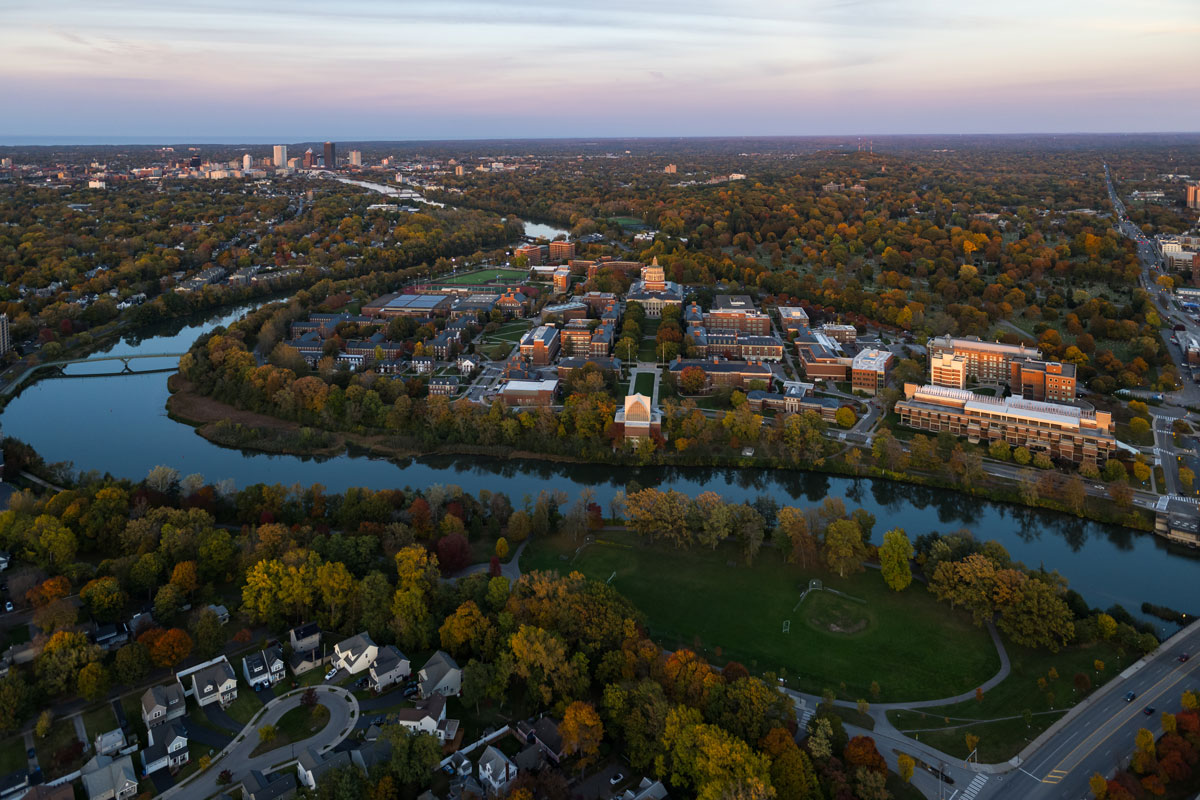 Aerial view of the sprawling University of Rochester campus bordered by a meandering river in the foreground, surrounded by autumn-colored trees. The opposite bank features suburban neighborhoods, and a distant skyline of city buildings is visible under a partly cloudy sky.
