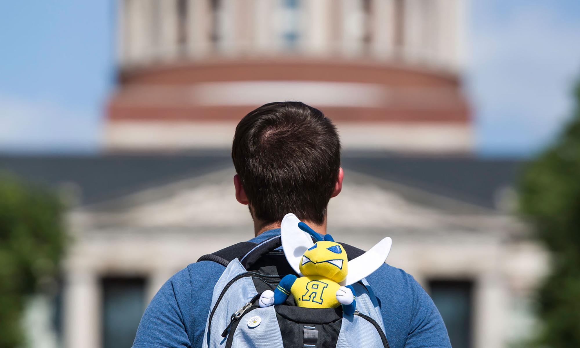 student facing Rush Rhees Library wears a backpack with a Rocky Yellowjacket mascot sticking out of it.